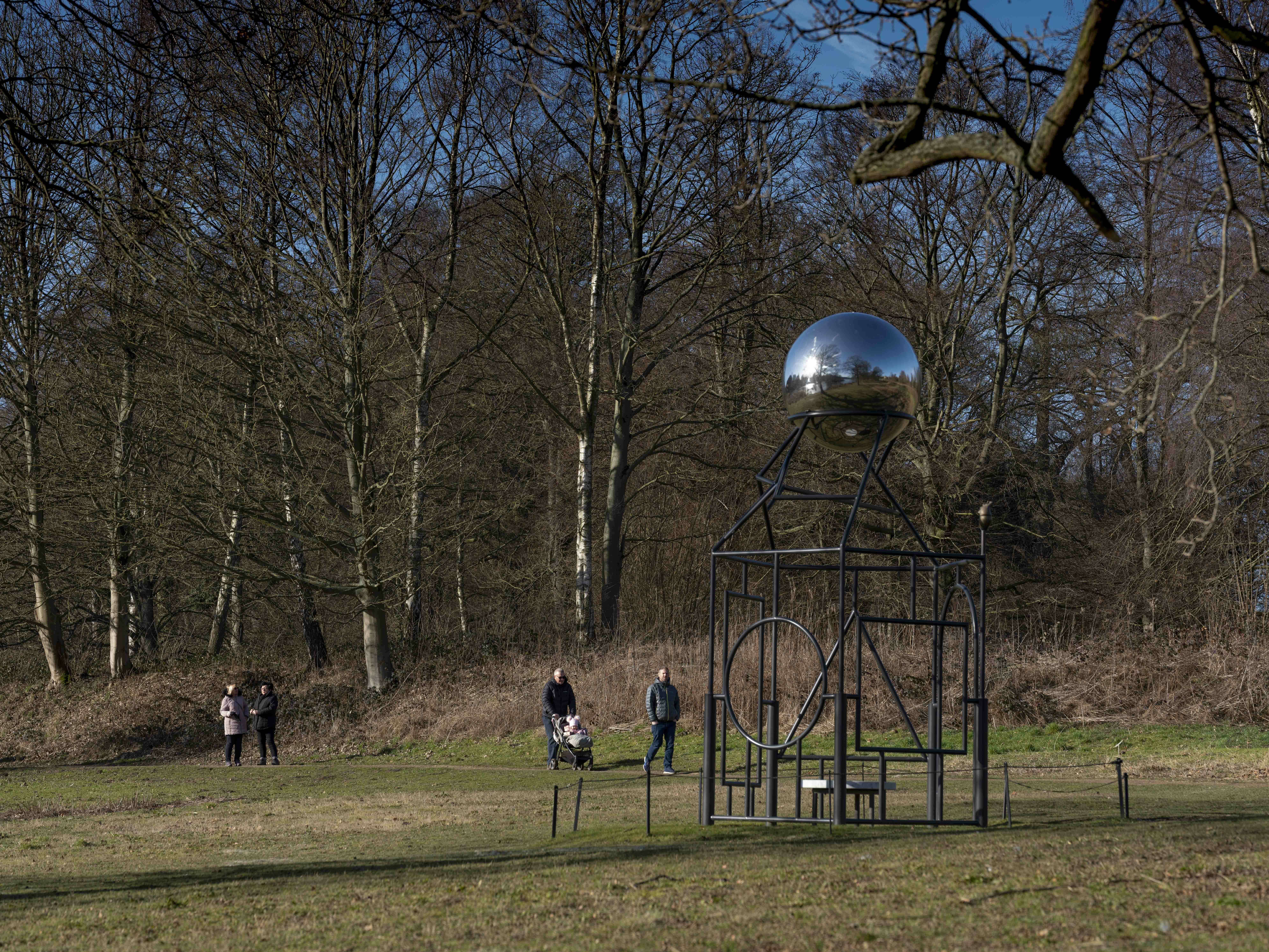 People with a pushchair walking past a metal frame sculpture with a silver ball at the top.