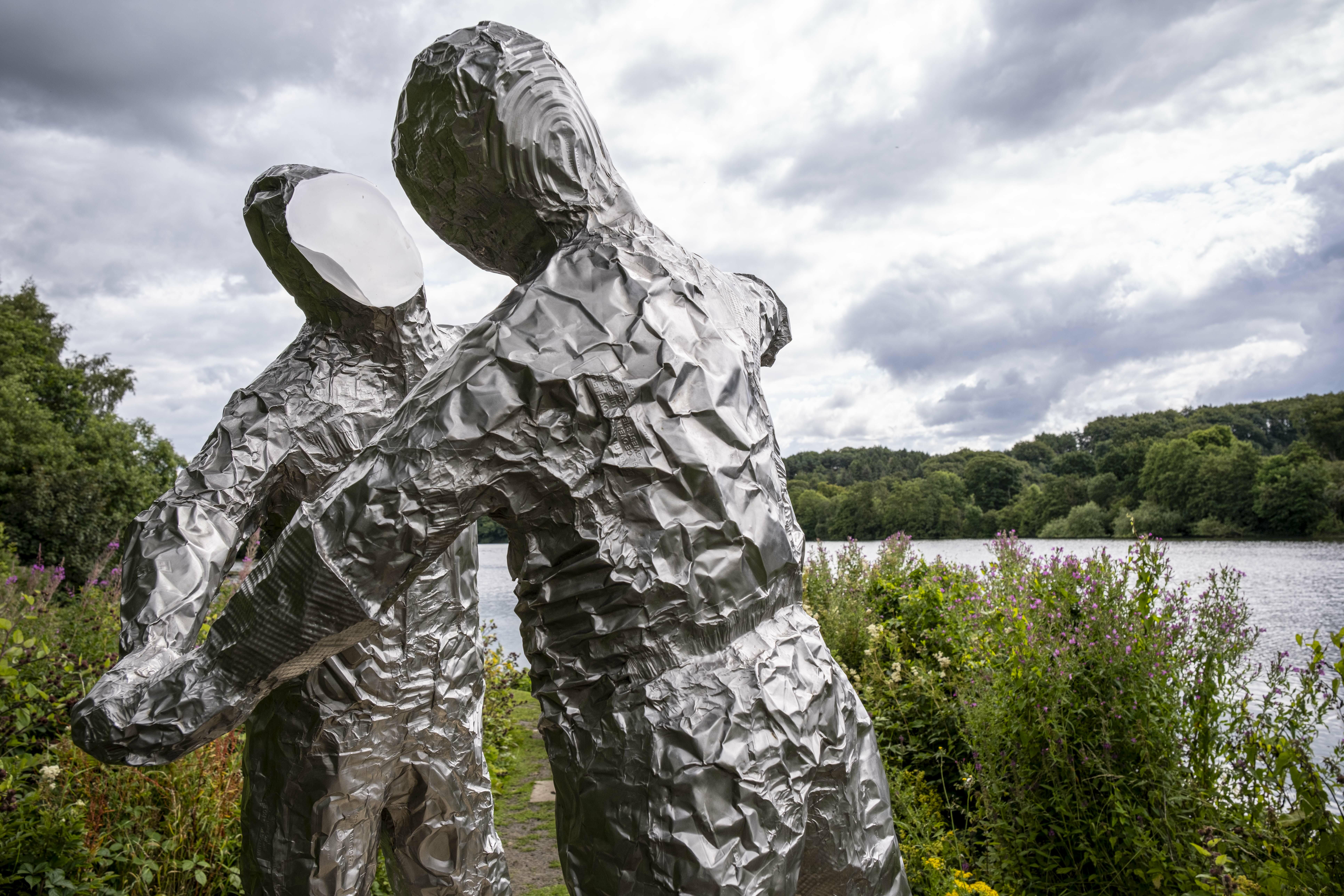Two silver figures in astronaut suits with flat expressionless faces, in an embrace. A lake is behind them.