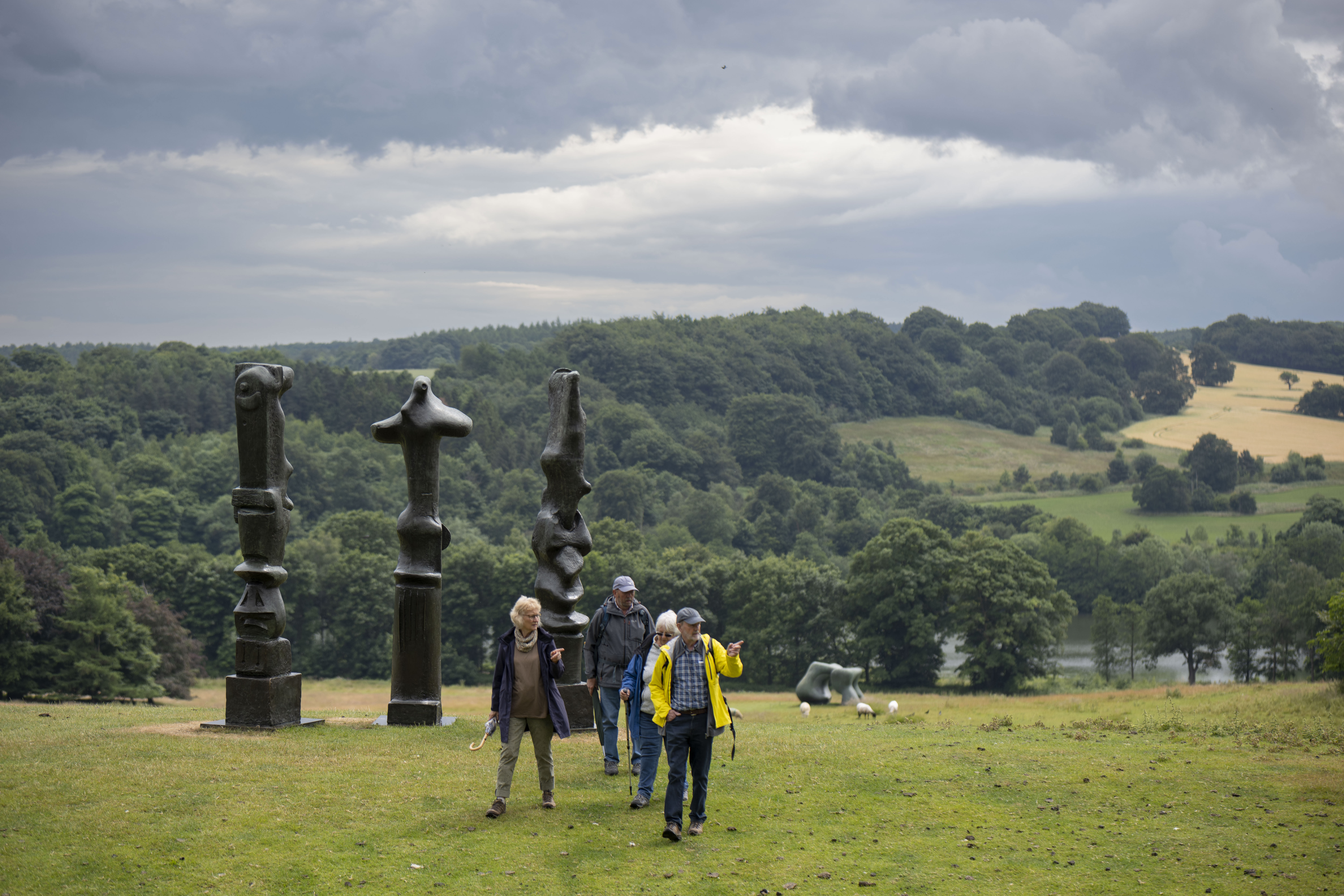 People in raincoats walking past three tall thin bronze sculptures looking out over the landscape