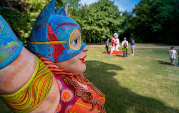 Close up of a human figure sculpture, with colourful clothing and Batman style mask. People looking at sculptures are in the distance.