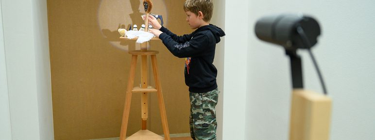A young boy stands at a wooden stool, focused on a small model illuminated by a circle of light. The scene is set in a minimalist room with a plain wall, and a directional light is visible in the foreground.