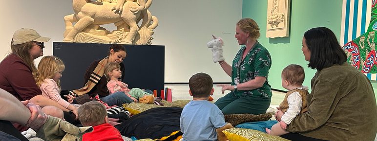 A group of children and adults sit on colourful cushions in an art gallery. A facilitator, wearing a green top, engages the children with a story, while a large sculptural artwork of a creature is displayed in the background.