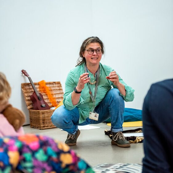 A storyteller crouches in front of a wooden box filled with colourful objects.