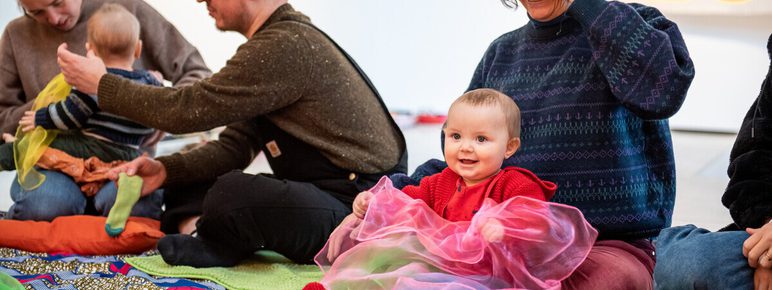 A group of adults and babies sit on colourful mats in an art gallery. A smiling baby in a red outfit plays with sheer, colourful fabrics, while an adult nearby watches. Others are engaged with their babies, creating a lively, creative atmosphere.