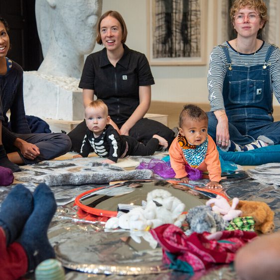 A group of adults and two babies sit on a reflective surface in an art space. The adults, dressed in casual clothing, engage with the children, who are exploring the area. Artworks are visible in the background, creating a creative atmosphere.