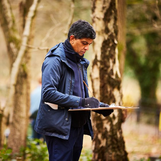 A man stands in a wooded area, focused on sketching on a clipboard.