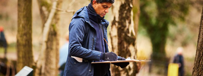 A man stands in a wooded area, focused on sketching on a clipboard.