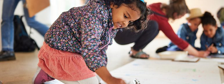 A young girl is kneeling on the floor, focused on drawing with crayons on a large sheet of paper. In the background, other children and adults are engaged in various art activities in a bright, spacious room.