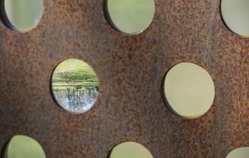 A close-up of a rusted metal panel featuring circular cut-outs. Through one of the holes, a reflection of a serene landscape with trees and grass is visible, contrasting with the rough texture of the metal surrounding it.