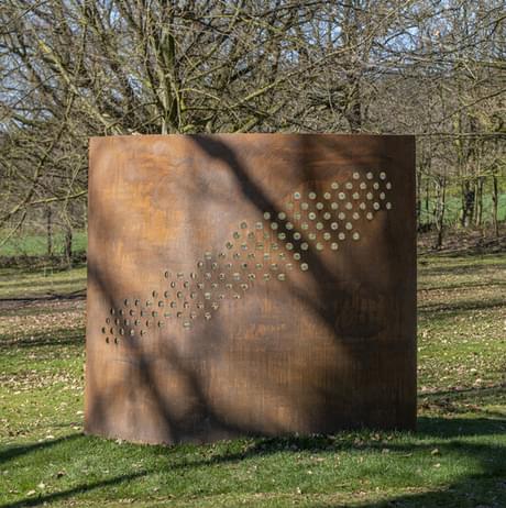 A large, rusted metal sculpture stands in a grassy area, featuring a pattern of circular cut-outs arranged in a diagonal line. Surrounding the sculpture are bare trees and a clear blue sky, creating a serene outdoor setting.