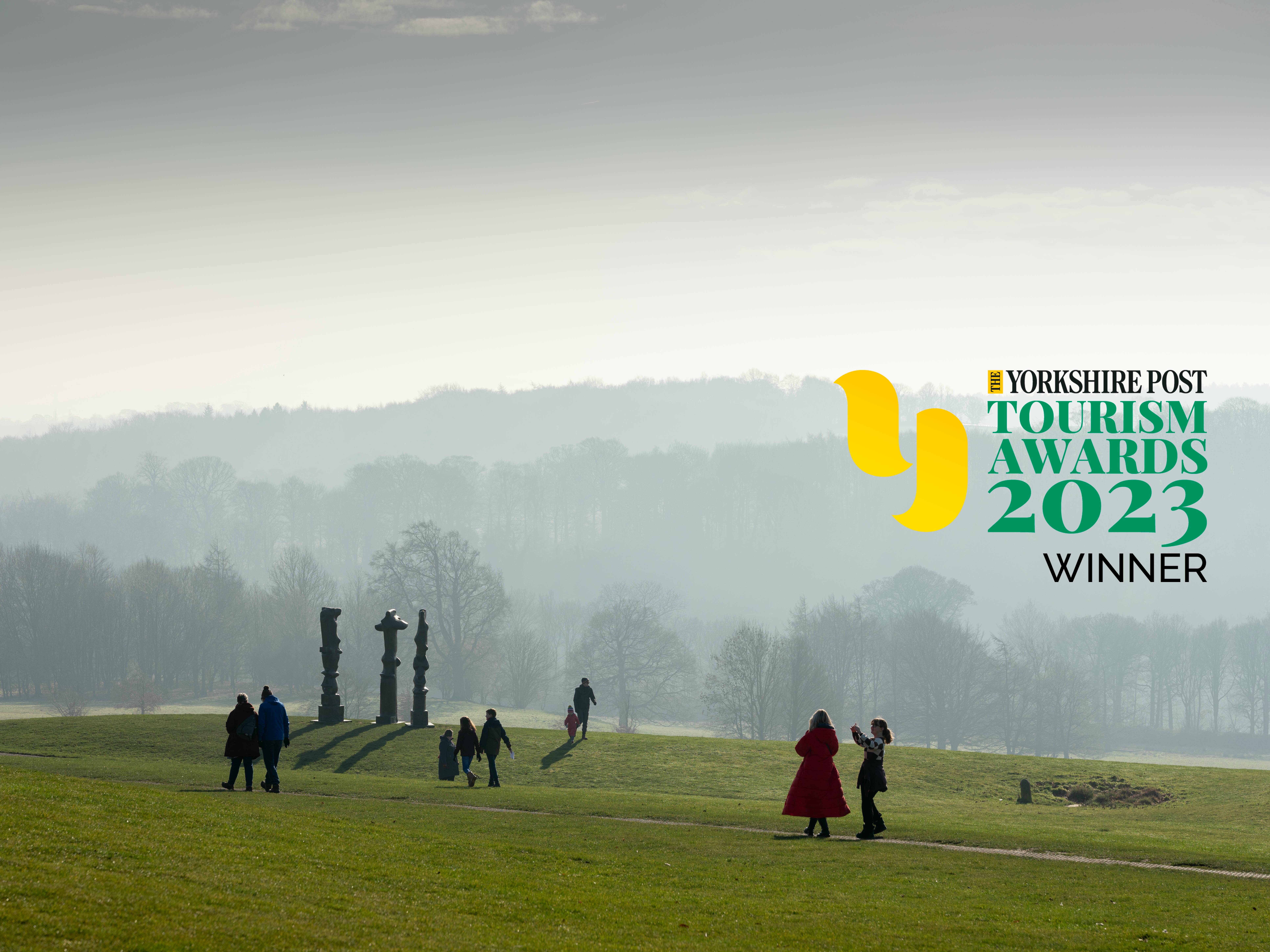 People walking across a hillside, with three tall thin silhouetted sculptures and trees in the background. The Yorkshire Post Tourism Award 2023 Winner logo is in the top right corner.