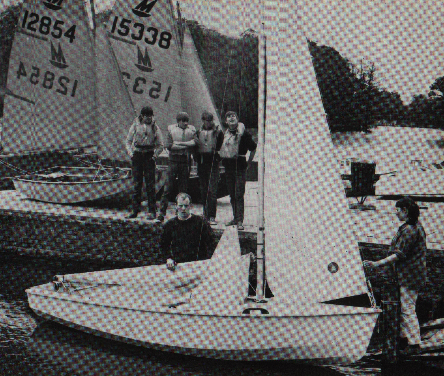 Four boys wait on the dock by the lake to get in to the sail boat held by two adults.