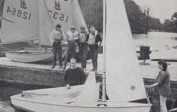 Four boys wait on the dock by the lake to get in to the sail boat held by two adults.