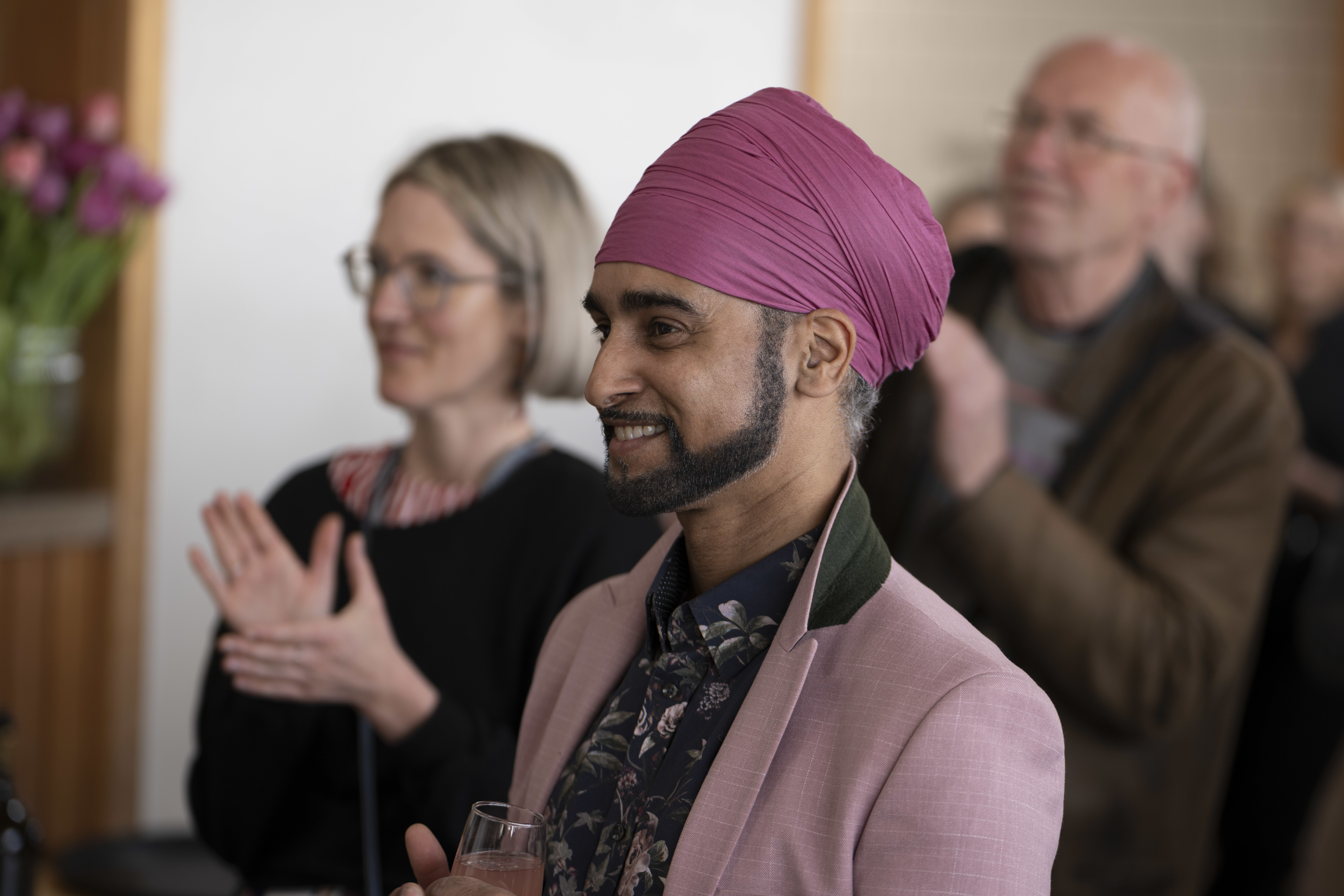 A man wearing a turban smiling with a champagne glass in his hand and a woman clapping behind him.