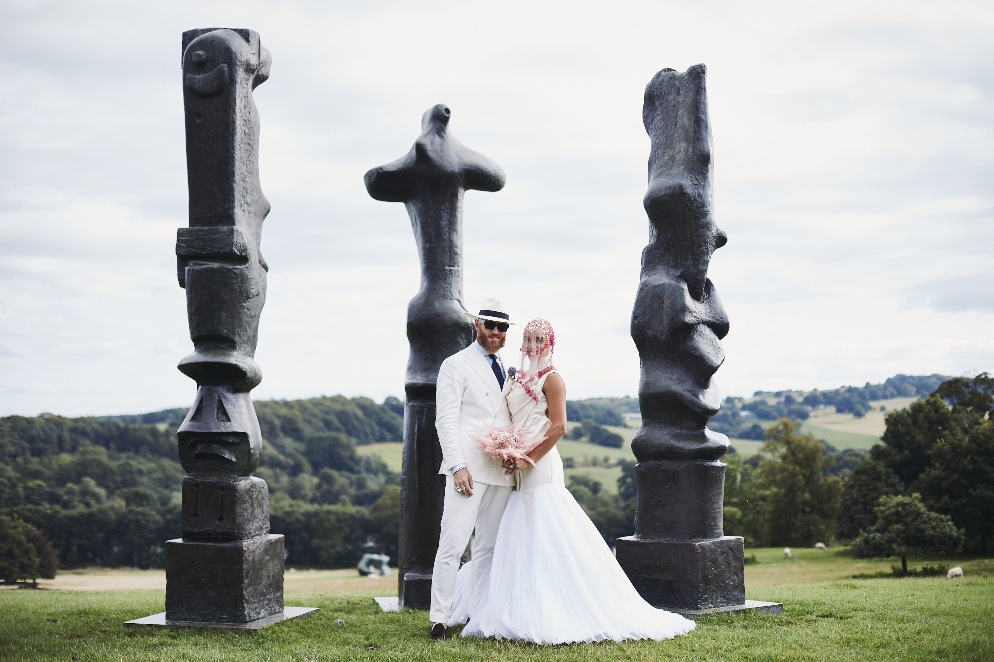 A bride and groom, wearing a long white dress and white suit, standing in front of three tall thin bronze sculptures outdoors