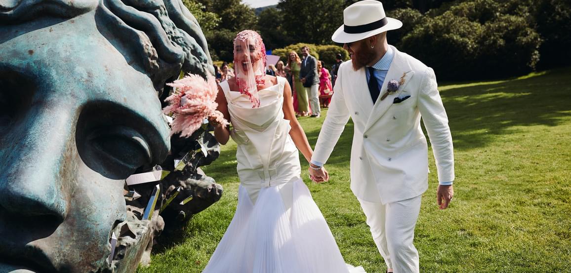 A bride and groom, wearing a white structured wedding dress and white suit, holding hands while walking past a giant bronze head sculpture.