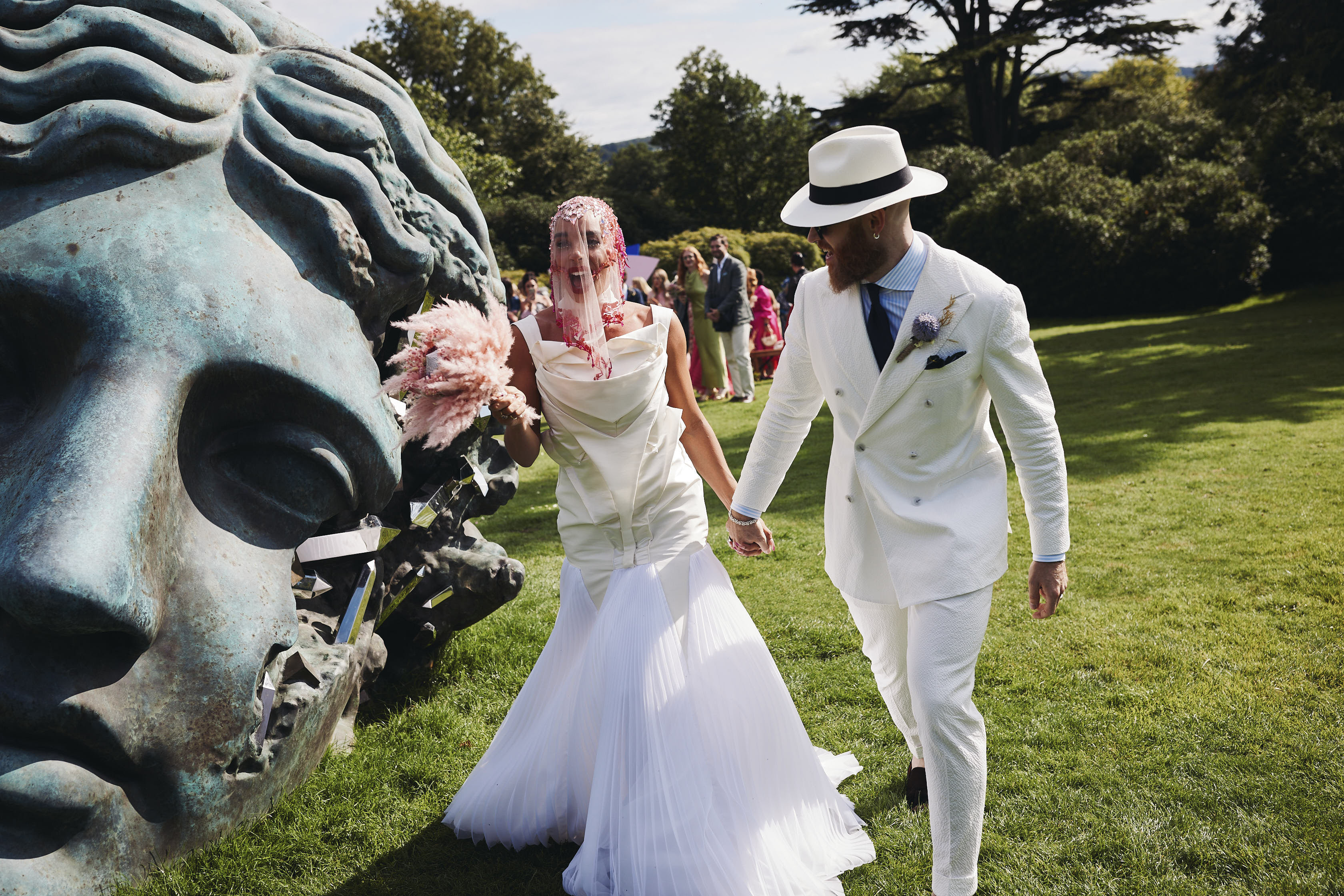 A bride and groom, wearing a white structured wedding dress and white suit, holding hands while walking past a giant bronze head sculpture.