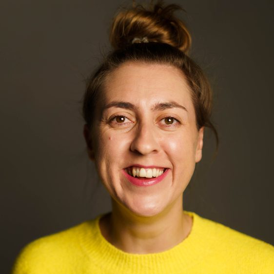 A smiling woman with brown hair in a bun, wearing a yellow sweatshirt