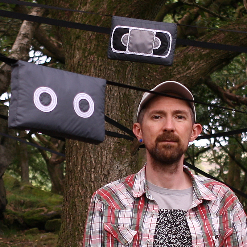 A man with a brown beard, wearing a baseball cap and checked shirt, standing in front of textile sculptures of cassette and VHS tapes suspended from a tree
