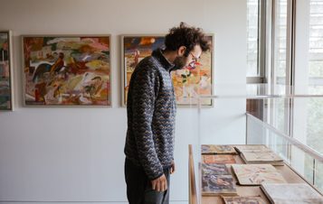 A man wearing a patterned fleece looks at a display of sketchbooks in a display case. Framed paintings are on the wall behind him.
