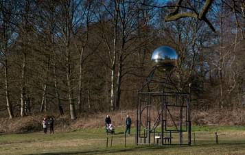 People with a pushchair walking past a metal frame sculpture with a silver ball at the top.