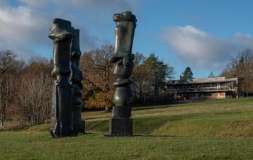 Three tall thin bronze sculptures with YSP Centre behind them