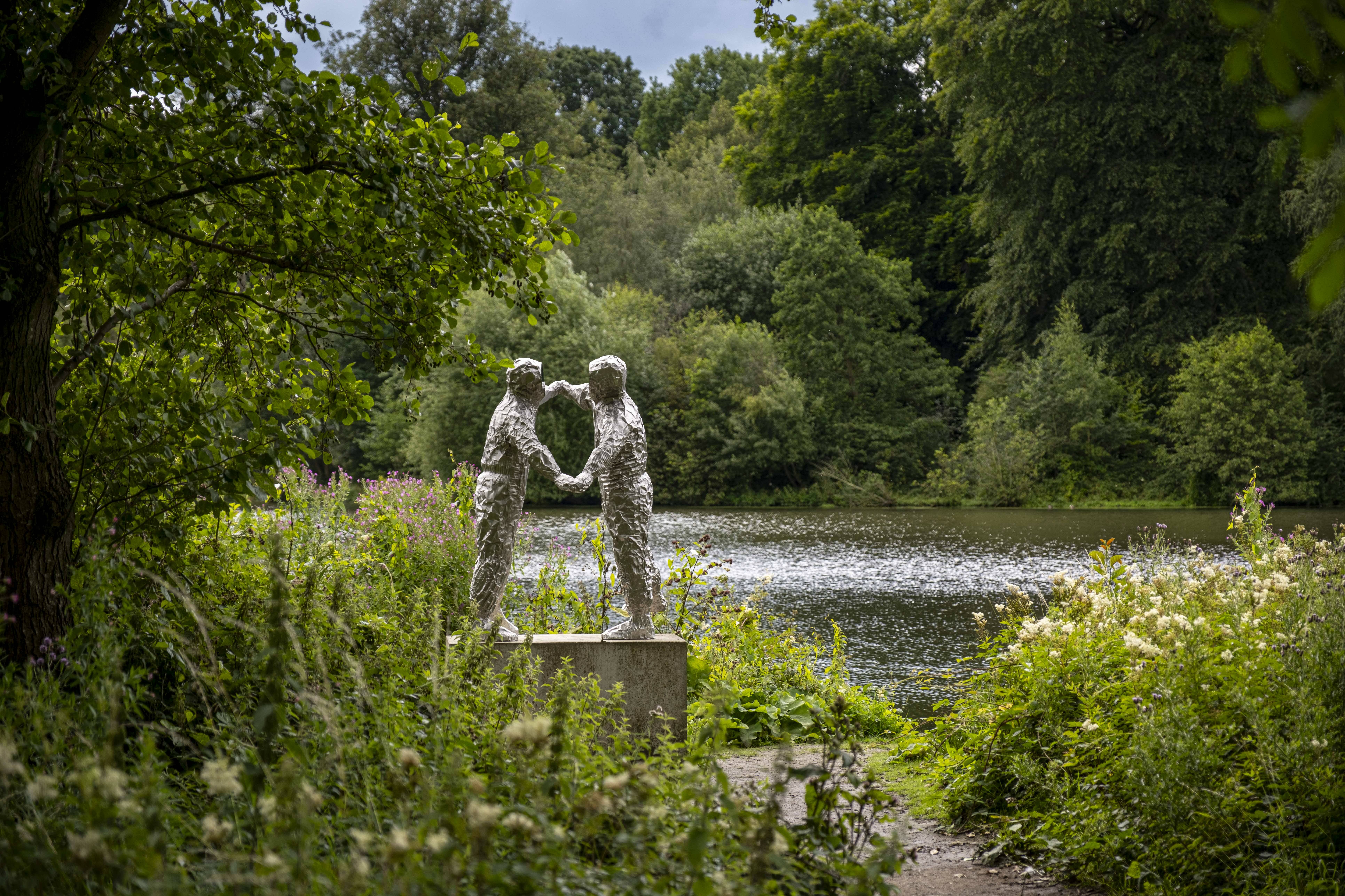 A silver sculpture of two figures dancing. A lake, trees and foliage surround it.