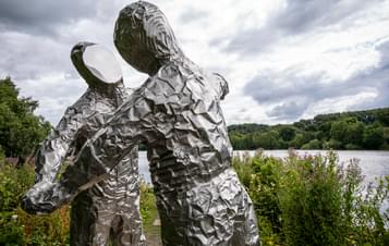 Two silver figures in astronaut suits with flat expressionless faces, in an embrace. A lake is behind them.