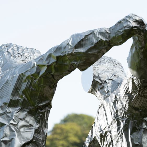 Two silver figures in astronaut suits with flat expressionless faces, in an embrace.