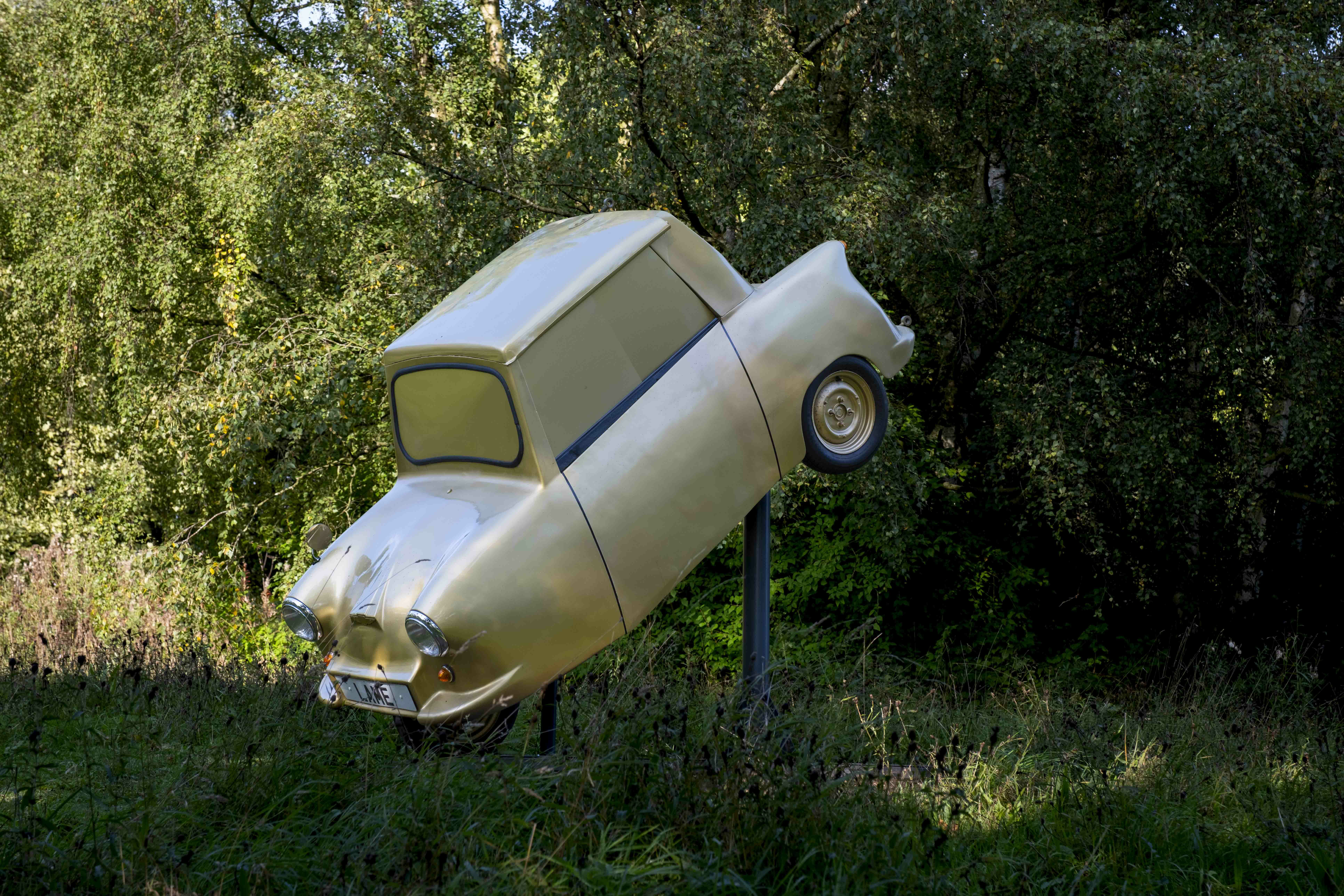 A small gold car suspended at a 45 degree angle with trees behind it.