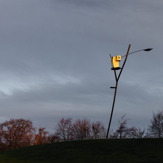 A tall, slender sculpture stands on a grassy hill, featuring a unique birdhouse design at its top. The sky is overcast with soft grey clouds.