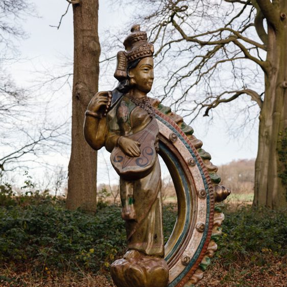A bronze sculpture of a human figure in traditional South Asian dress, with half replaced by a decorative curved section. It is displayed outdoors in woodland.