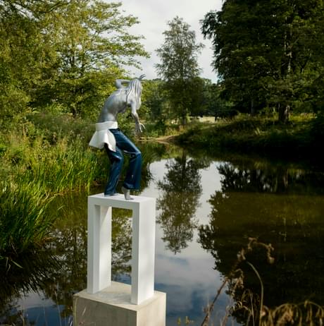 A female figure wearing jeans standing on a white plinth at the edge of a lake, surrounded by foliage