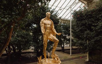 A gilded gold sculpture of a human figure with the skin removed revealing the musculature, displayed inside a glass house with large bushes. The figure is holding a knife and scissors and standing on a circular table.