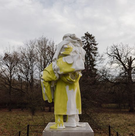An abstract sculpture of a humanoid figure covered in draped fabric, displayed outdoors surrounded by trees.
