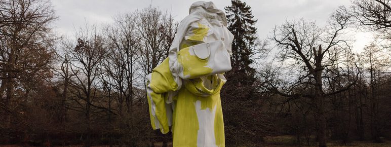 An abstract sculpture of a humanoid figure covered in draped fabric, displayed outdoors surrounded by trees.