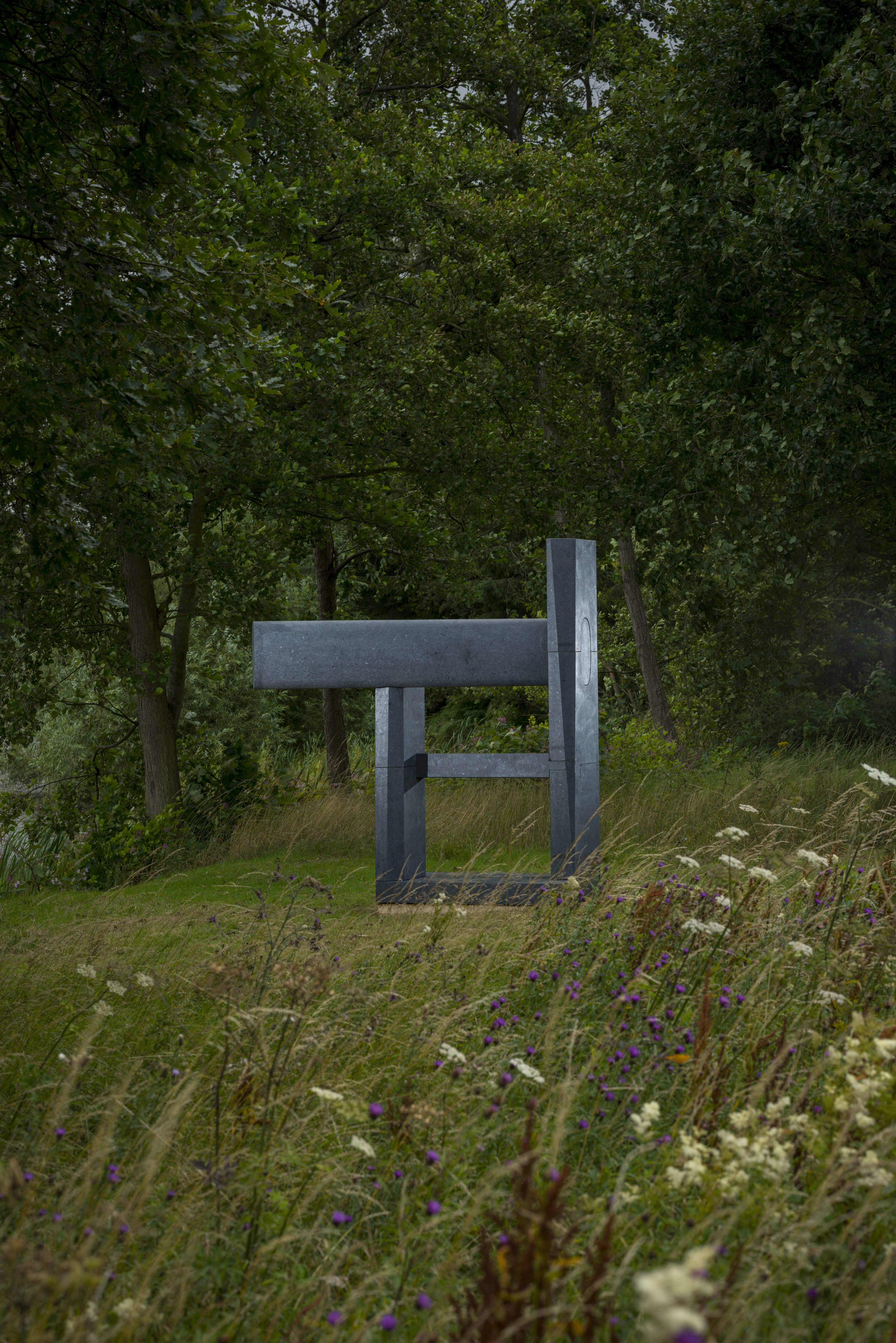 A dark grey abstract sculpture next to a lake, surrounded by wild flowers