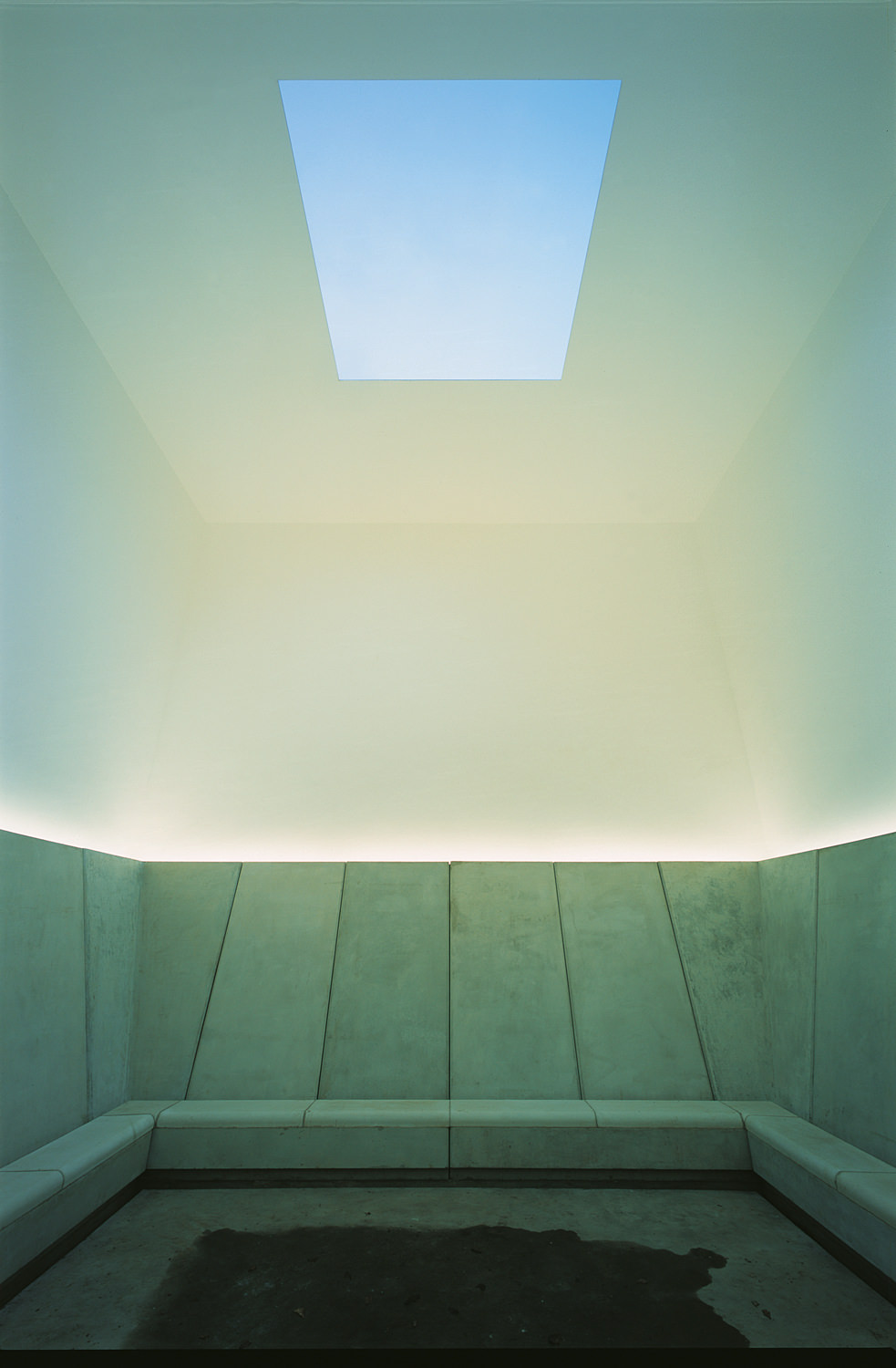 A room with concrete bench seating and a square hole cut into the ceiling revealing the sky