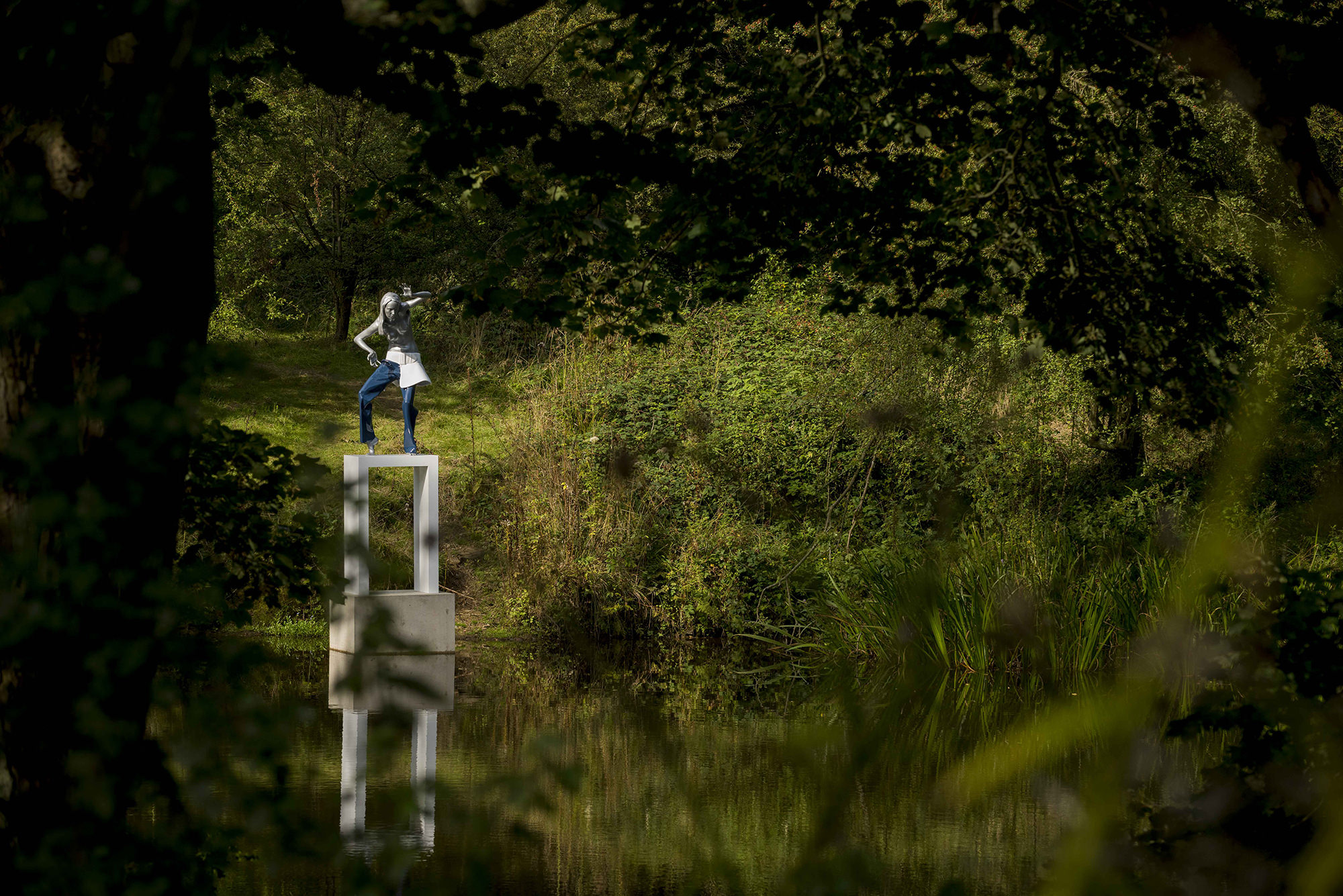 A female figure wearing jeans standing on a white plinth at the edge of a lake, surrounded by foliage