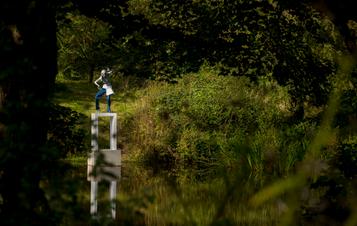 A female figure wearing jeans standing on a white plinth at the edge of a lake, surrounded by foliage