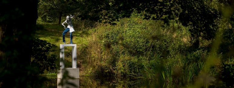 A female figure wearing jeans standing on a white plinth at the edge of a lake, surrounded by foliage