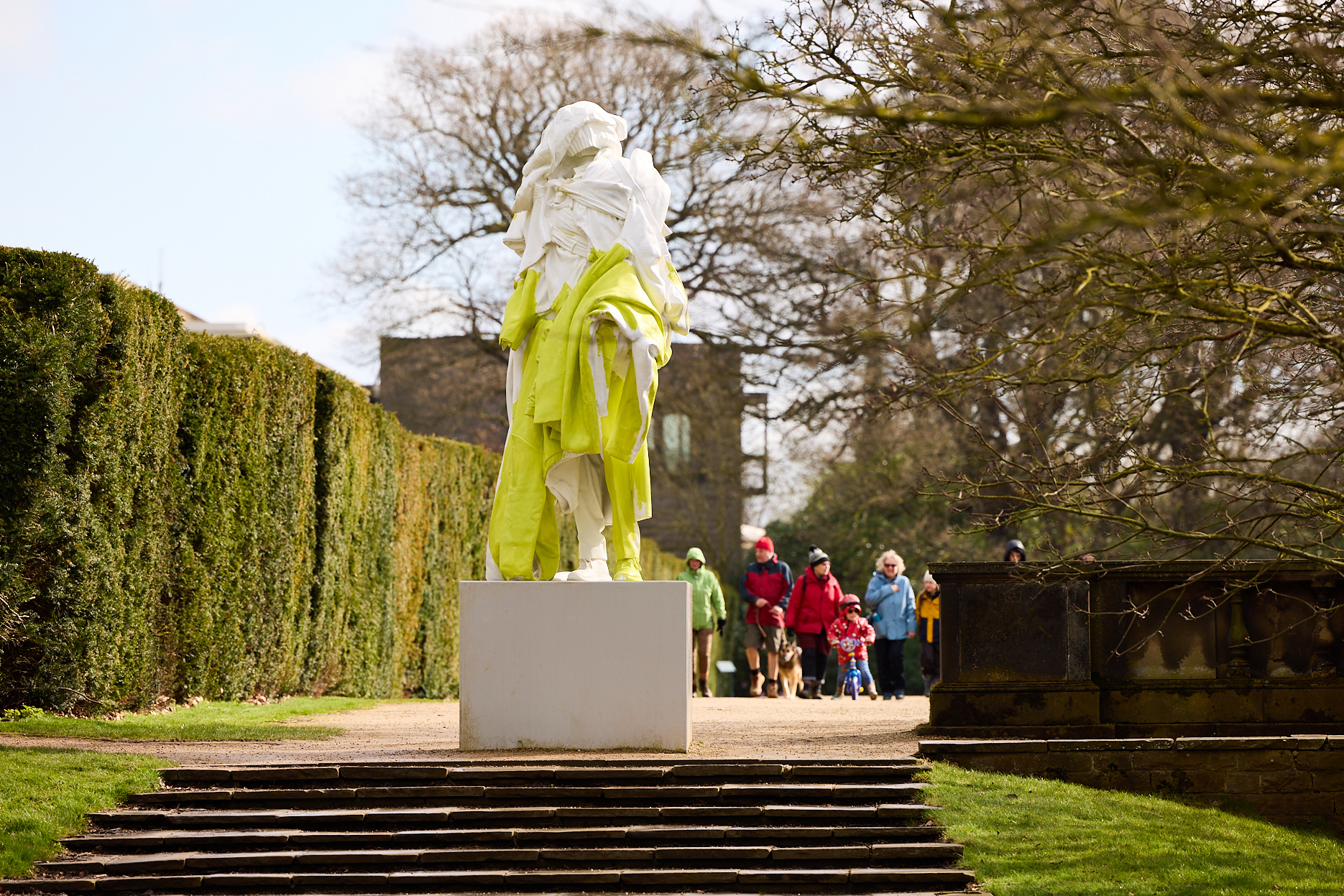 A group of people in colourful coats walking towards a yellow and white sculpture of a figure draped in clothing