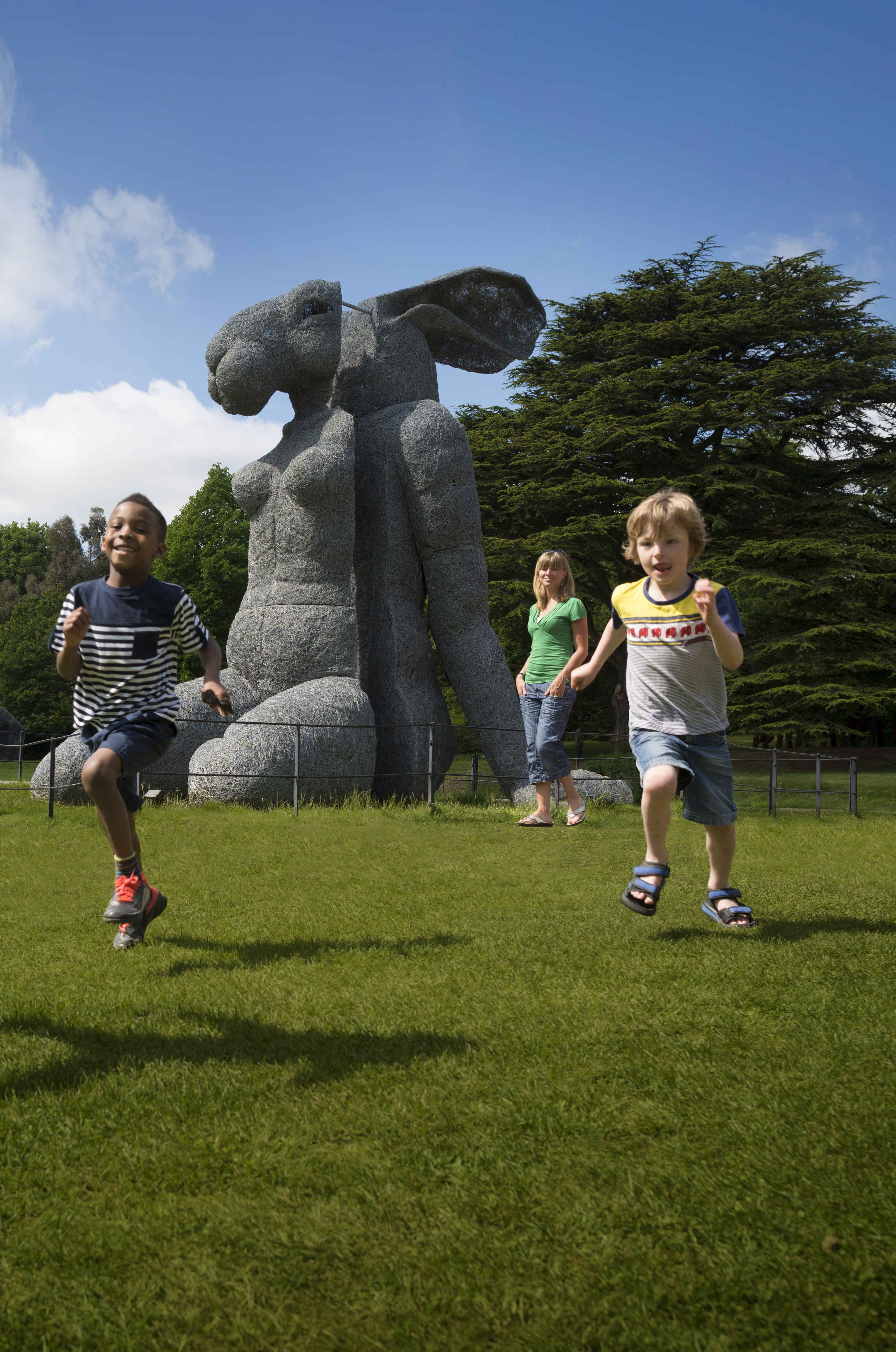 A group of children running in front of a large sculpture of a female body with a rabbit head.