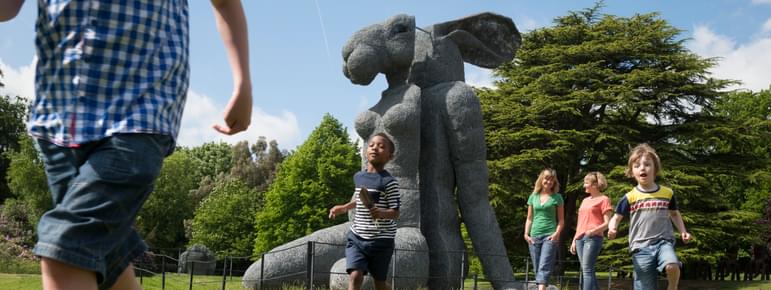 A group of children running in front of a large sculpture of a female body with a rabbit head.