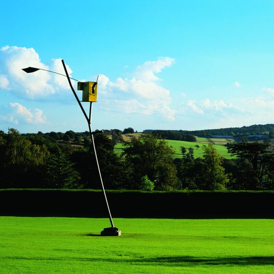 A tall sculpture of a watchtower with a yellow abstract shape at the top of a long tilted pole, displayed outdoors.