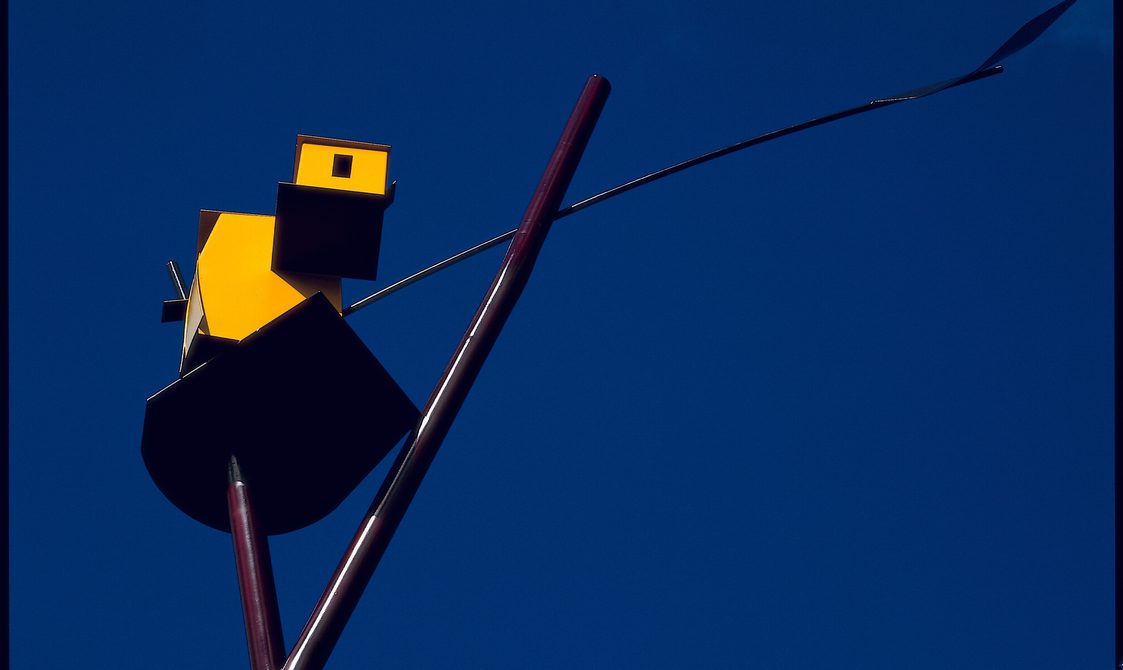 Detail of a sculpture depicting an abstract yellow structure perched at the top of a thin metal pole, set against a vibrant blue sky