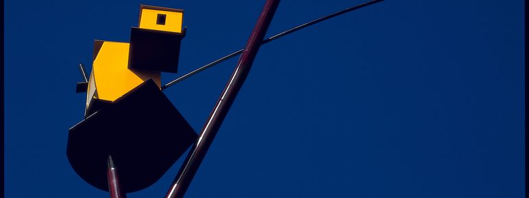 Detail of a sculpture depicting an abstract yellow structure perched at the top of a thin metal pole, set against a vibrant blue sky
