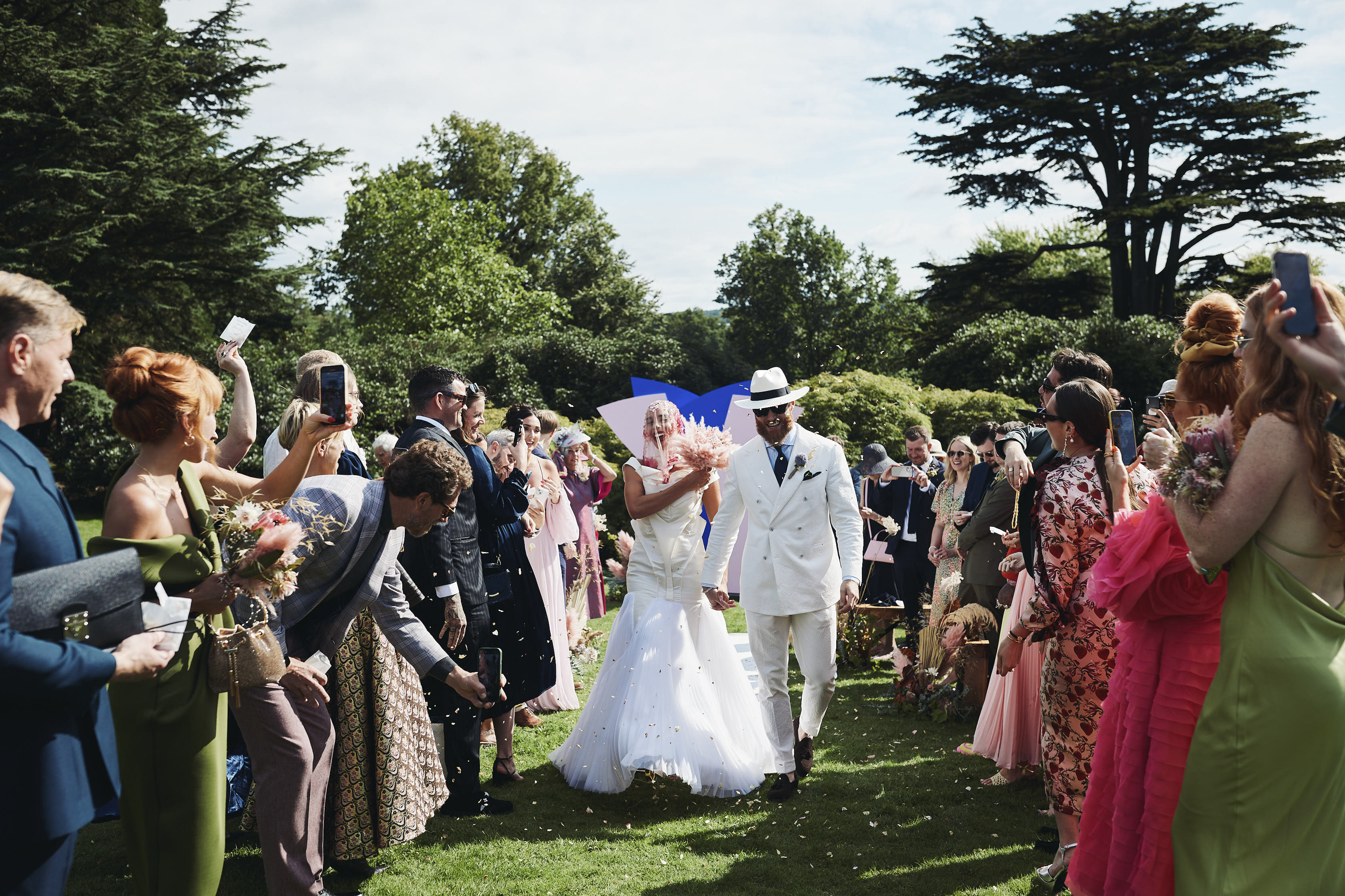 A bride and groom, wearing a long white dress and white suit, walking down an outdoor aisle surrounded by their guests.