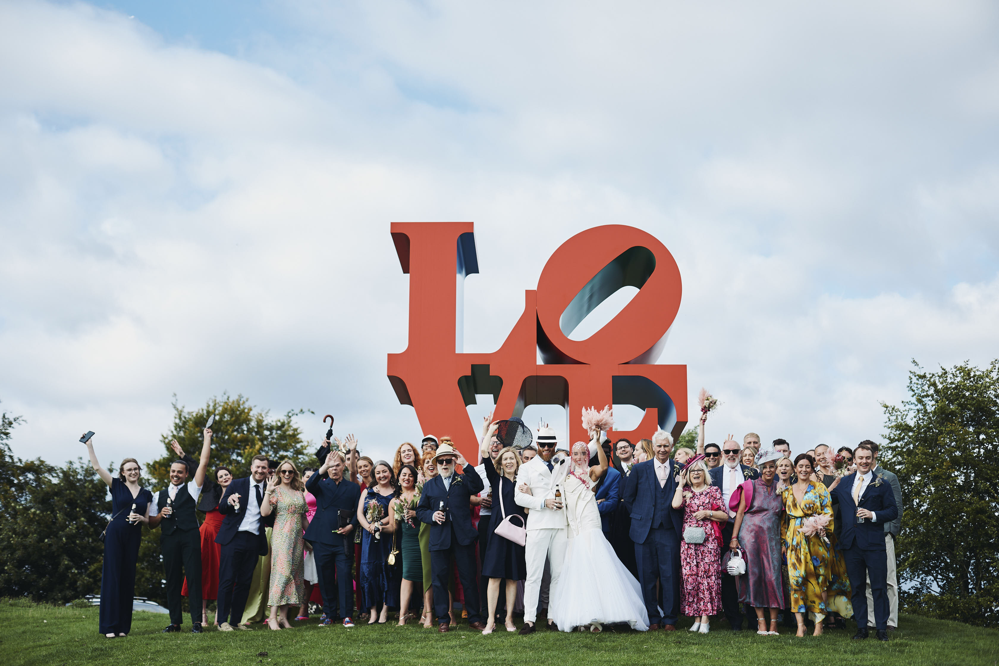 A wedding party, including the bride and groom and guests, standing in front of a large red LOVE sculpture outdoors.