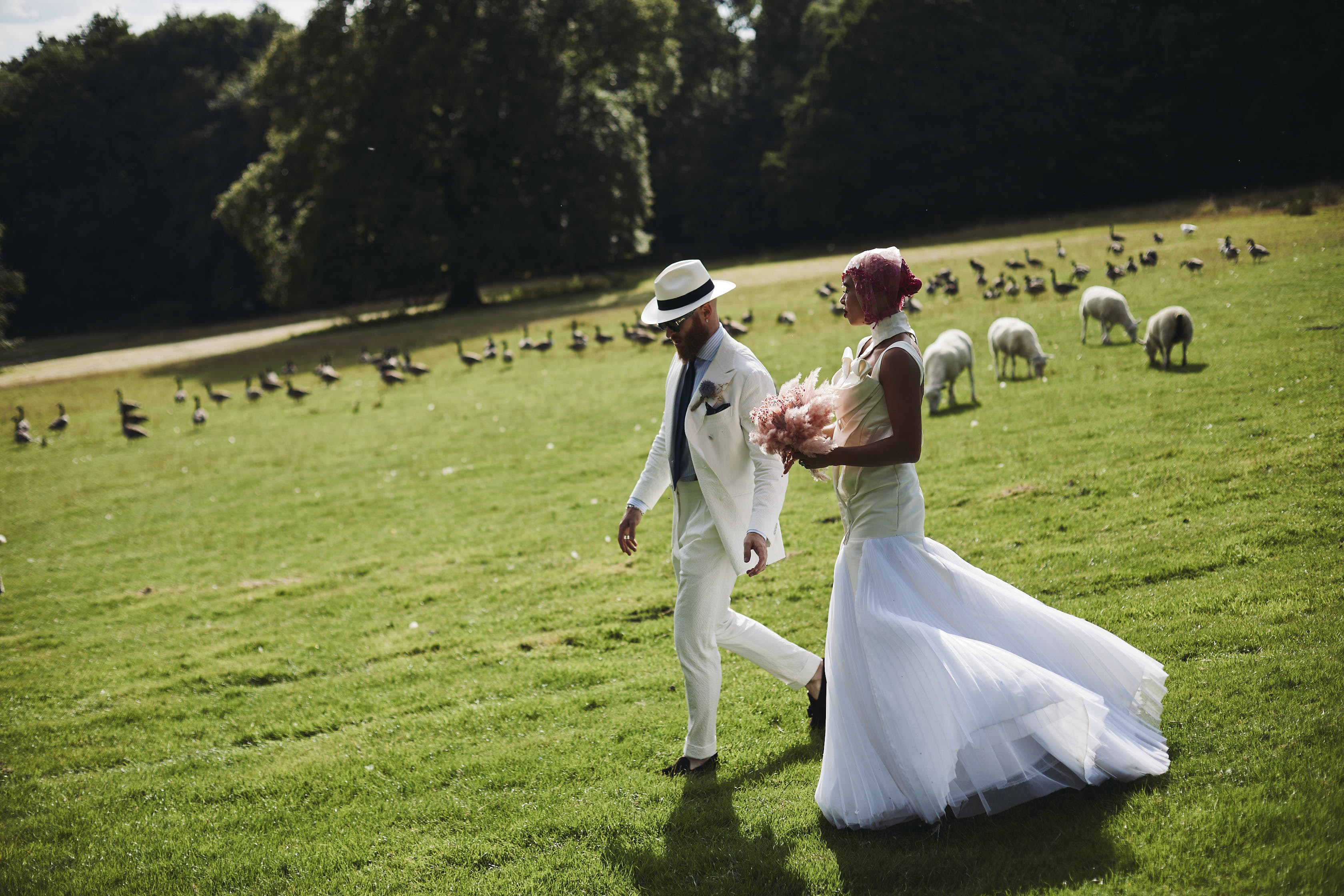A bride and groom, wearing a long white dress and white suit, walking outdoors through a field of sheep and geese